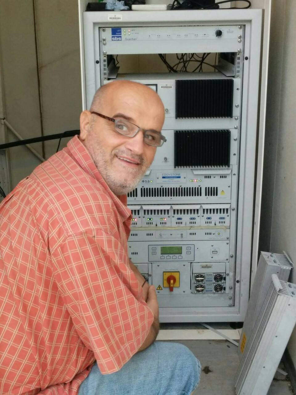 Zejnullah Osmani kneeling beside an FM transmitter rack with Eddystone broadcast amplifiers